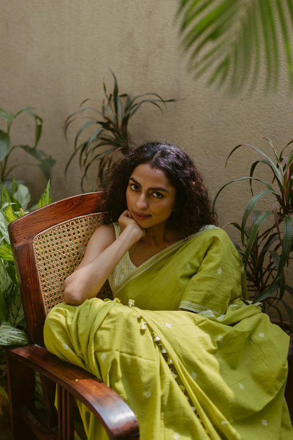 Woman wearing lime green and white handloom cotton Jamdani saree sitting in a garden.