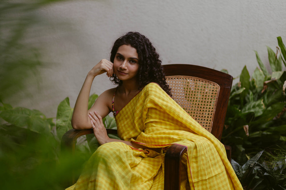 Curly-haired woman in a yellow checked handloom cotton saree, seated on a cane chair amid greenery, from the Varsaa Edit Checks & Plaids collection.